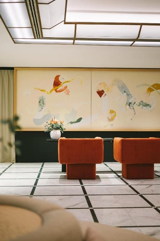 Two orange armchairs face a wall with abstract art and a black console table holding a white vase with flowers; the floor features a geometric tile pattern.